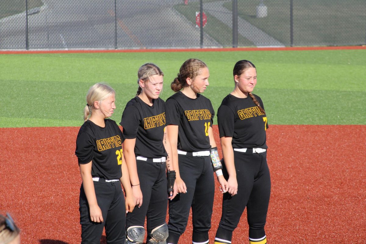 While the flags are presented for the National Anthem, varsity players Haidyn Cleveland (10), Tessa Jensen (11), Kendall Johnson (10), and Amelia Fliege (10) stand at the pitchers mound holding hands.