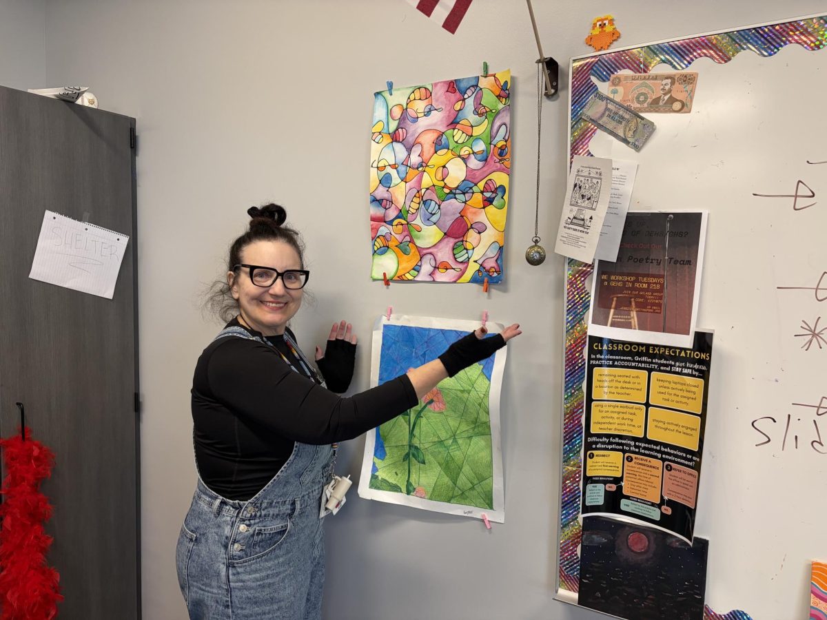Posing for a photo, English teacher Jennifer Smith stands in front of student artwork, an old pocketwatch and an Iraqi dollar. Almost all of Smith's classroom decorations are student artifacts.